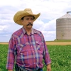  Onaldo veste uma camisa xadrez, usa um bigode e veste chapéu. Ao fundo está a sua plantação. 